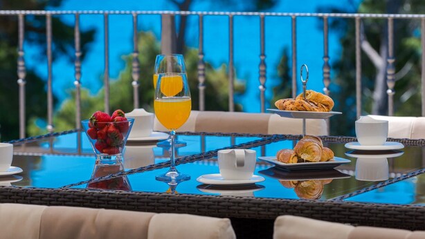 A breakfast table with a glass of orange juice, strawberries, croissants, and a white cup on a glass table with a view of palm trees. - Spacious 5 bedroom Stone Villa with Private Pool and Sea view  (Mlini)