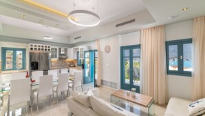 A modern kitchen with a central island, a dining area with a glass table and chairs, and a living area with a sofa and a coffee table. - Royal Villa Pedi - Symi (Symi)