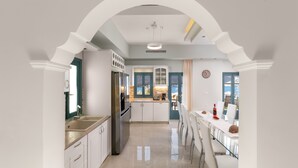 A modern kitchen with white cabinetry, a stainless steel refrigerator, and a dining area with a glass table and chairs. - Royal Villa Pedi - Symi (Symi)