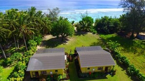 Dos edificios amarillos con techos grises, rodeados de exuberante vegetación y palmeras, con vista a una playa y el océano al fondo.