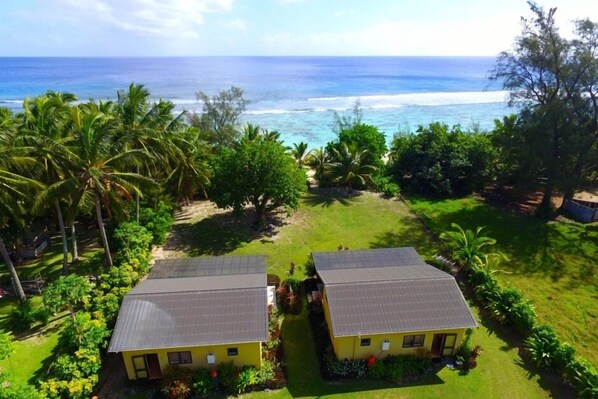 Two yellow buildings with grey roofs, surrounded by lush greenery and palm trees, overlooking a beach with the ocean in the background.