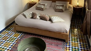 Un dormitorio con cama, almohadas, mesita de noche con lámpara, un adorno en la pared y suelo estampado.