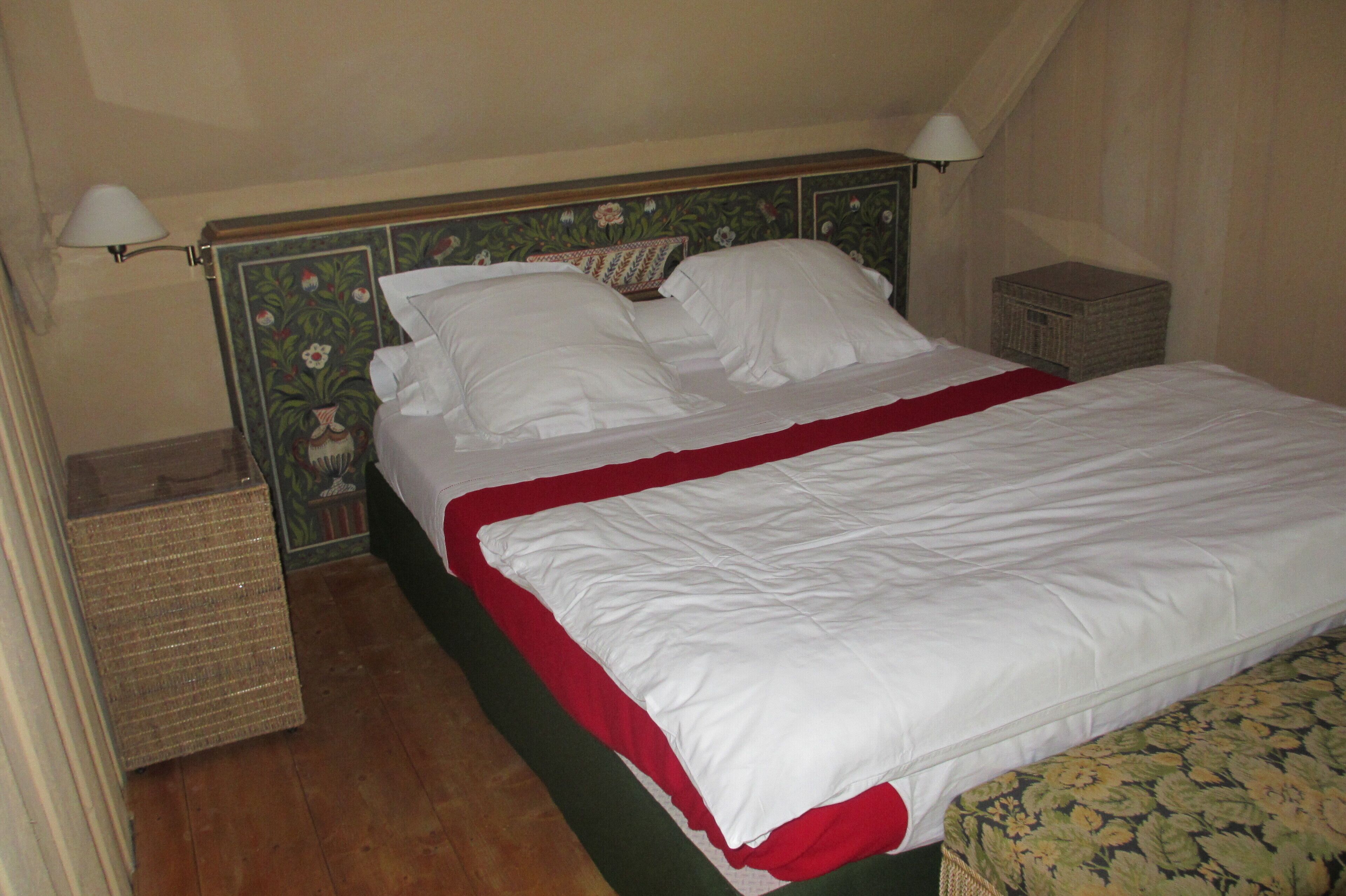 Un letto ben rifatto con lenzuola bianche e un tocco di rosso, una testiera in legno con motivi floreali e un comodino con una lampada.