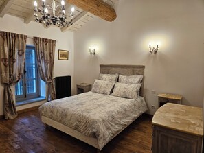Une chambre à coucher avec un lit en bois, un lustre, deux appliques murales, une fenêtre avec des rideaux et un radiateur.