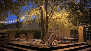 Une terrasse extérieure avec des chaises longues et une table, entourée d’arbres et illuminée de guirlandes lumineuses.