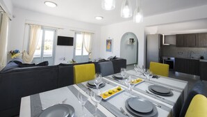 A modern dining area with a long table set for a meal, a sofa, and a kitchen in the background. - 3 bedroom Luxury villa , 200m by  famous Monolithos  beach!  (Έξω Γιαλός Καρτεραδου)