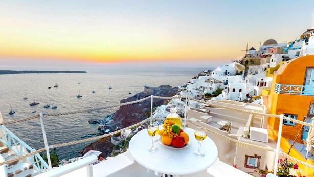 Blick auf das Meer mit einem Tisch, der mit Obst und Weingläsern gedeckt ist, mit Blick auf eine Küstenstadt mit weißen Gebäuden und Booten auf dem Wasser.