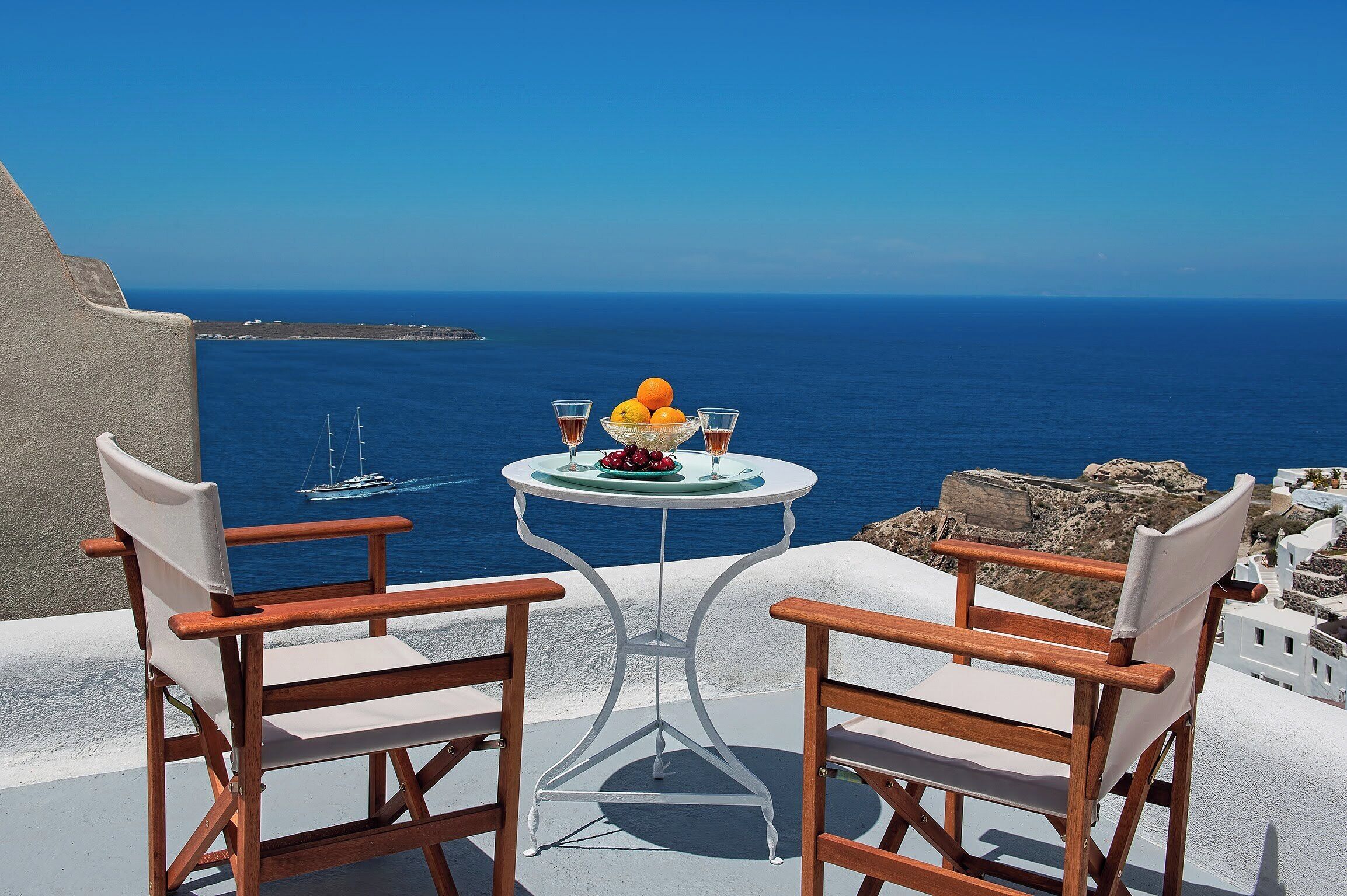 A table with a bowl of fruit and two glasses, overlooking a coastal view with a clear blue sky.