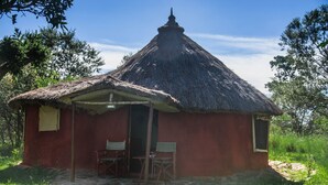 A small, single-story building with a thatched roof and a red exterior. There is a door leading to an interior space with a table and two chairs.
