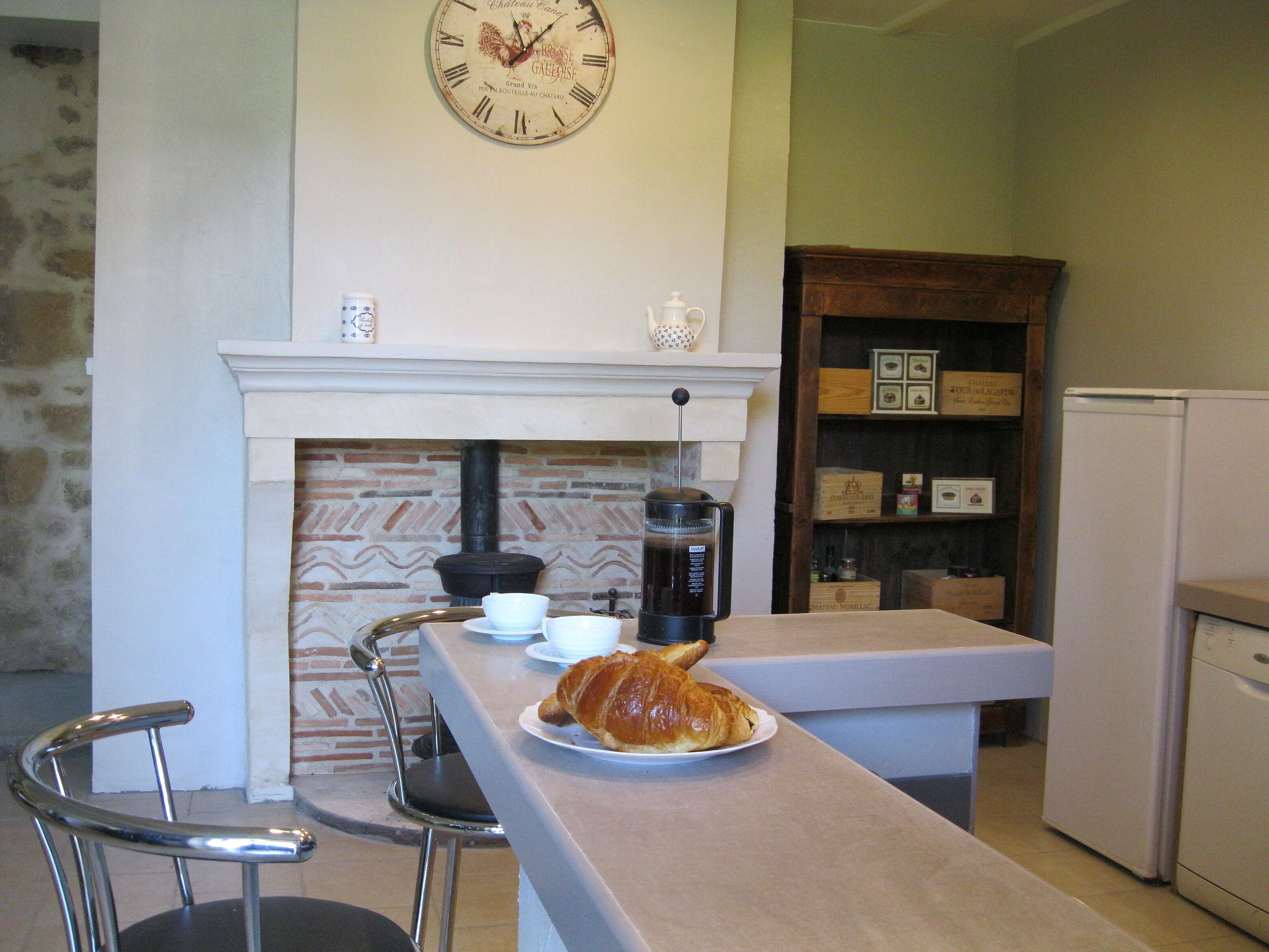 A kitchen with a stone wall, a clock, a coffee maker, a plate with a croissant, and a bar stool.