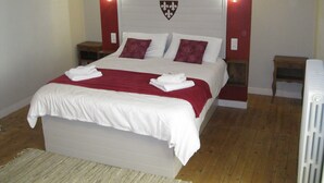 Un lit bien fait, avec du linge de lit blanc et un plaid rouge, deux tables de chevet en bois avec des lampes, et un mur d’accent rouge orné d’un emblème héraldique.