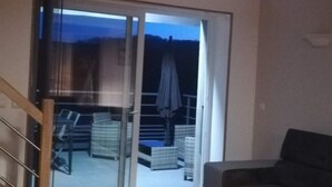 Een woonkamer met een donkerbank, een klein rond tafeltje en een schuifdeur die leidt naar een buitenruimte met rieten meubels en een terrasparasol.