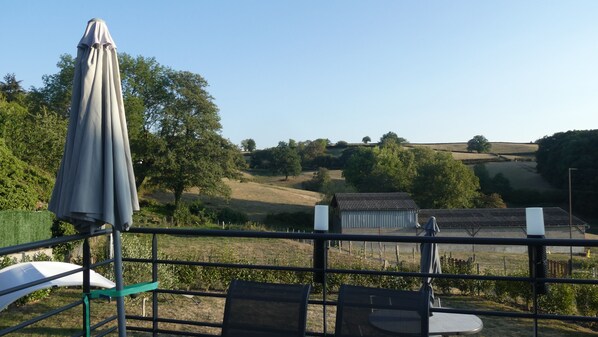 A terrace with a view of rolling hills and trees. There are chairs and an umbrella.