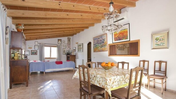 A dining area with a wooden table, chairs, and a fruit bowl. There is a bed with a blue cover in the background. - FINCA ES JARDI, pool, free internet WIFI, air conditioning, 2 bedrooms, 1 bathroom (Santanyí)