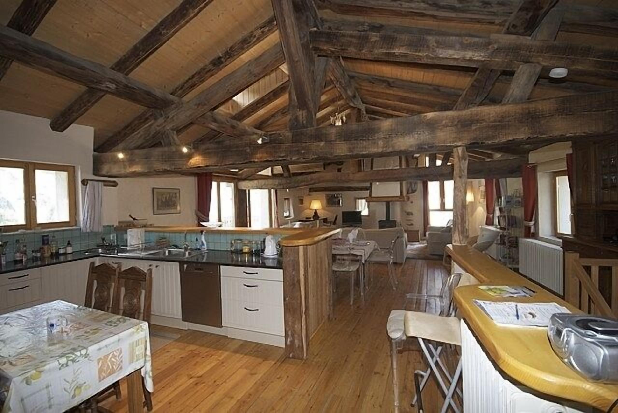 Une cuisine spacieuse avec des poutres en bois, un îlot central et un coin repas avec des chaises.