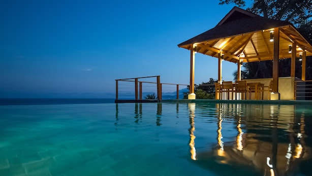An infinity pool with clear water reflecting lights, a wooden gazebo with seating, and a view of the ocean and mountains in the background. - Cliff House with Private Infinity Pool. 3 Minute Walk to the Beach. (Puntarenas)