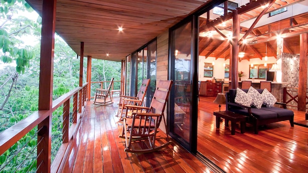 Una terraza de madera con mecedoras y un sofá, ventanas amplias y vistas al verdor.
