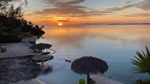 Une vue sur la plage avec un coucher de soleil, une piscine et un parasol en chaume.