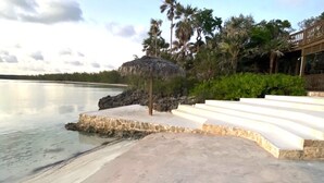 Une zone située en bord de mer, avec un parasol en chaume, des marches menant à la plage et des palmiers en arrière-plan.