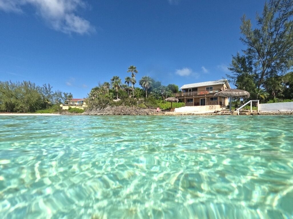 A beachfront property with clear water, a thatched-roof structure, and a large building with balconies.