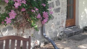 Un bâtiment rustique en pierre, doté d’une porte en bois et d’une fenêtre, ainsi que d’un banc et d’un arbuste en fleurs.