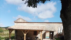 Un bâtiment rustique en pierre, doté d’un toit en tuiles, d’un patio couvert et d’une allée en gravier.