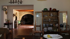 A dining area with a wooden table, chairs, and a plate with fruit. There is a bookshelf with various items and a clock on the wall.