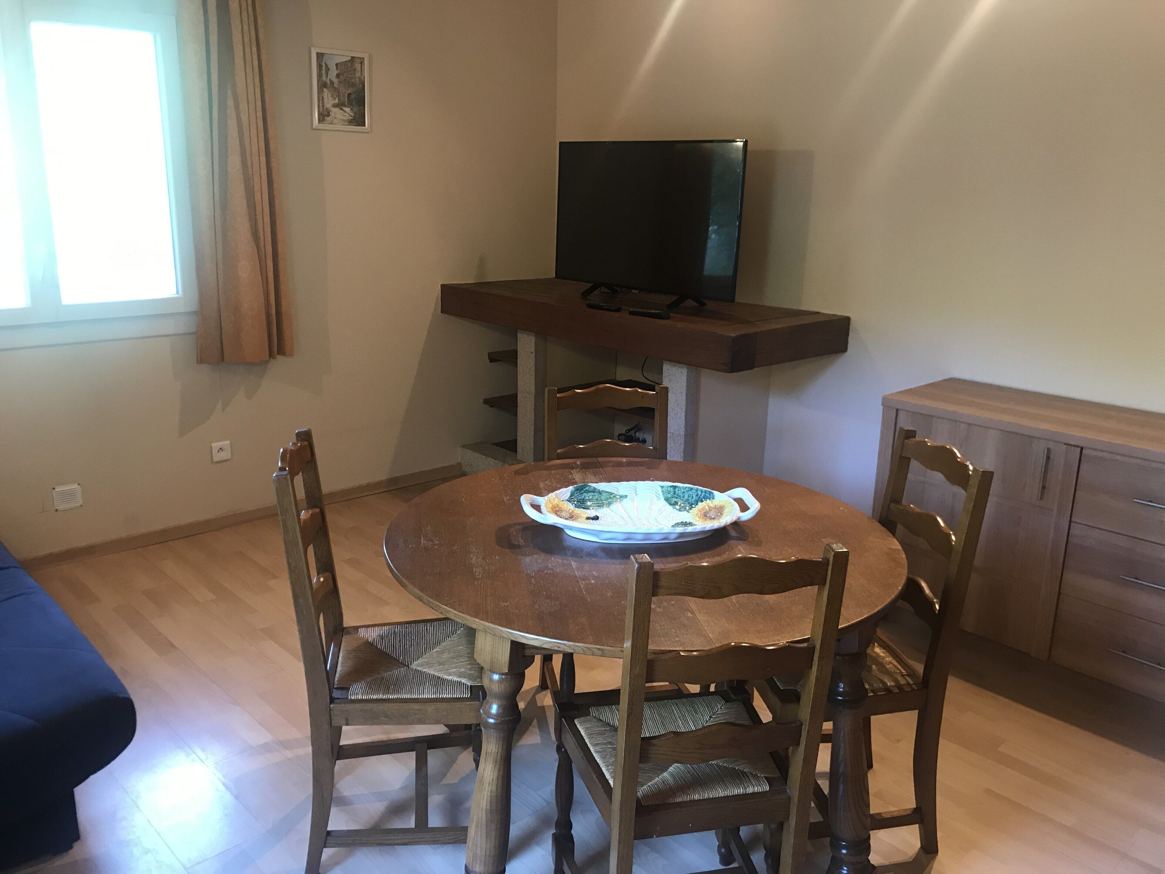 A dining area with a round wooden table, four chairs, a television on a stand, and a window with curtains.