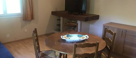 A dining area with a round wooden table, four chairs, a television on a stand, and a window with curtains.