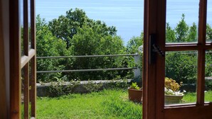 A view through a wooden door with a glass panel, revealing a lush green garden, a stone wall, and a clear view of the sea and distant mountains. - EPOUSTOUFLANTE SEA VIEW BREATHTAKING SEA VIEW8 people.9e pers possible (erbalunga)
