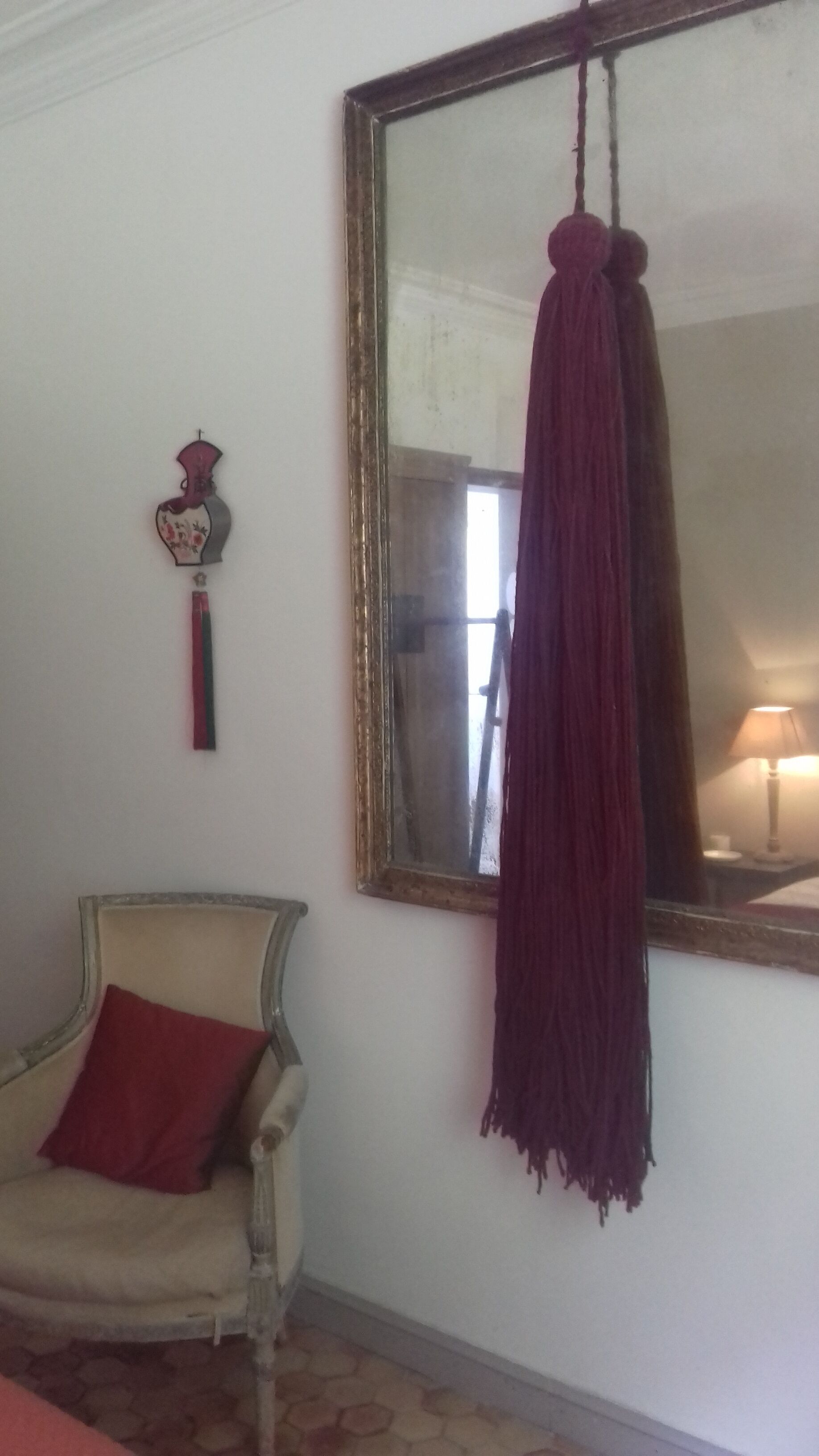 Een kamer met een grote spiegel, een hangende rode stof, een decoratieve vaas en een stoel met een rood kussen.
