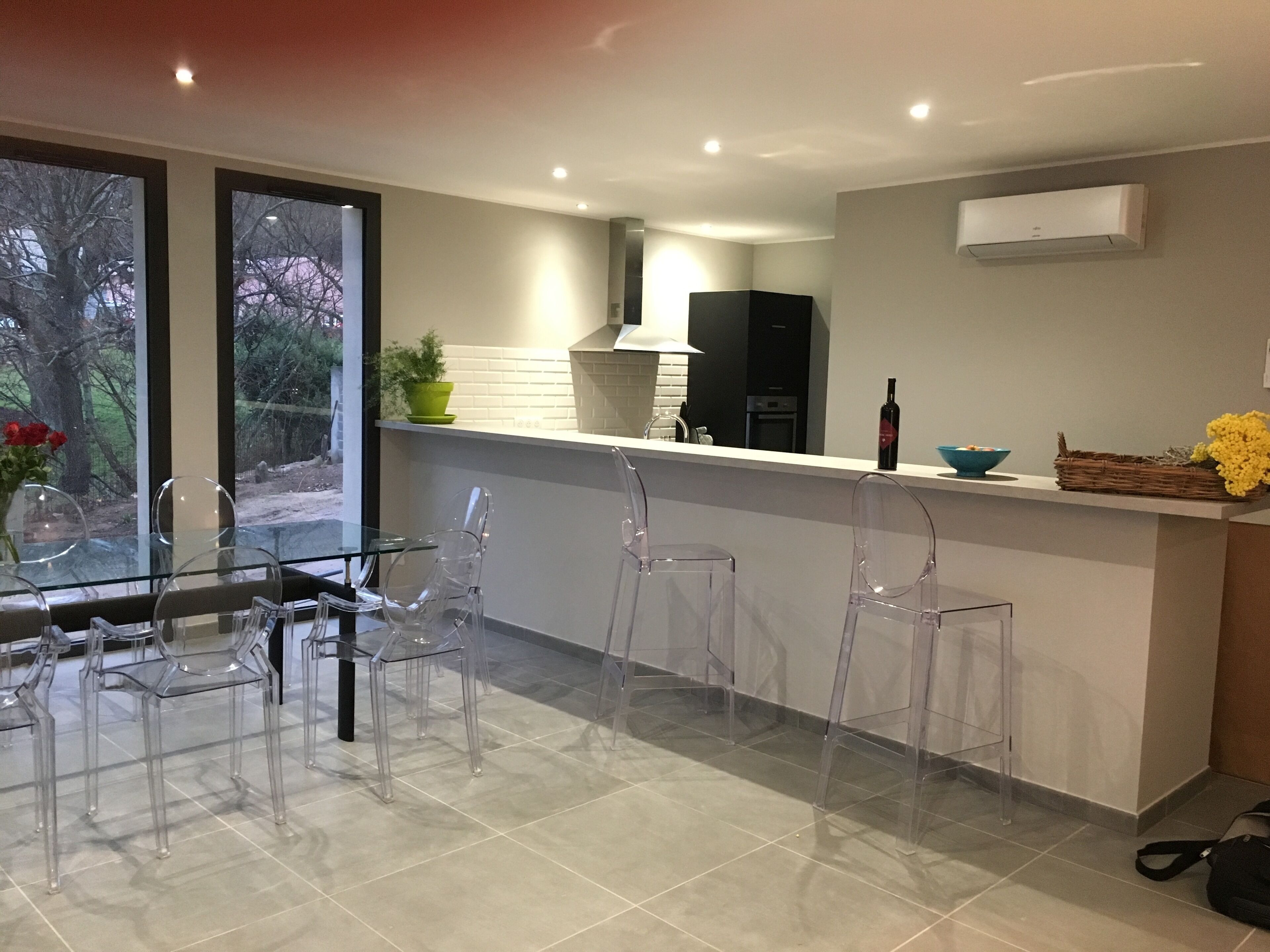 Een moderne keuken met een glazen eettafel en stoelen, een inbouwfornuis en een raam met uitzicht op een tuin.