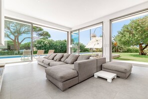 A modern living room with a sectional sofa, a coffee table, and large windows offering a view of a pool and garden. - Villa California - 6 bedrooms, swimming pool with lap lane and beach access on foot (porticcio)