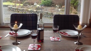 A dining table set with plates, glasses, and a bottle, overlooking a garden with trees and a stone wall.