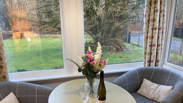 A room with a round table, two grey sofas, a vase with flowers, and a bottle of wine.