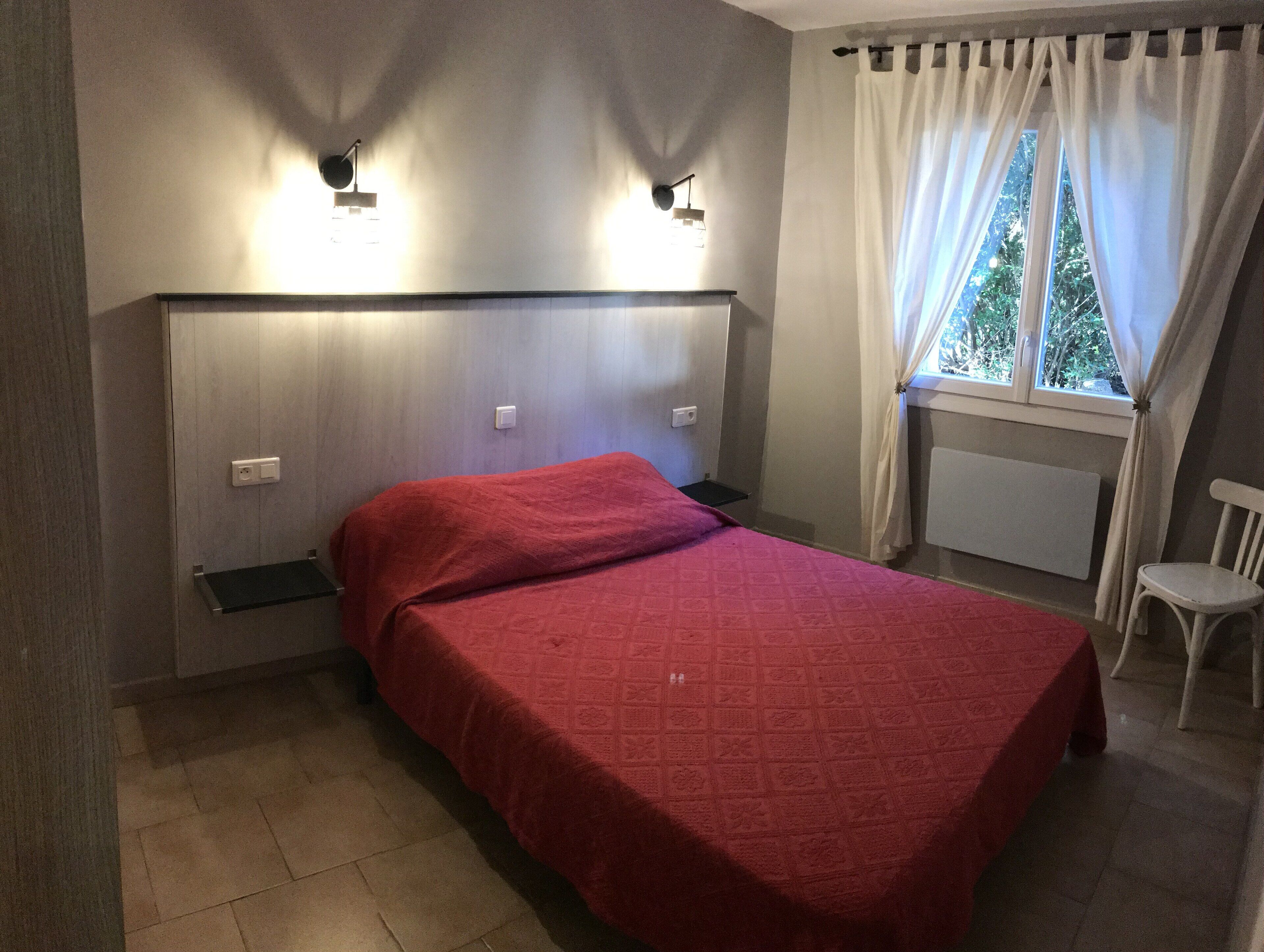 A bedroom with a bed, a nightstand, a window with curtains, and wall-mounted lights.