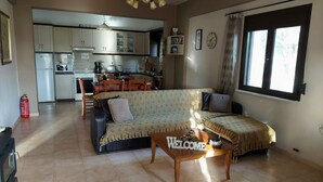 A living room with a sofa, a coffee table, and a kitchen in the background. - Villa Elia is situated in a quiet area, surrounded by olive trees with a private (Πιτσίδια)