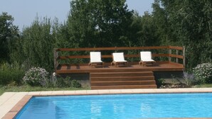 A swimming pool with three white lounge chairs on a wooden deck, surrounded by greenery.