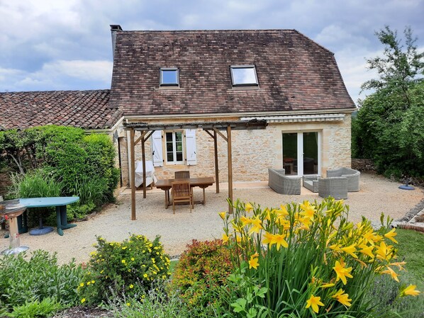 A stone house with a tiled roof, a covered patio with a table and chairs, and a garden with yellow flowers.