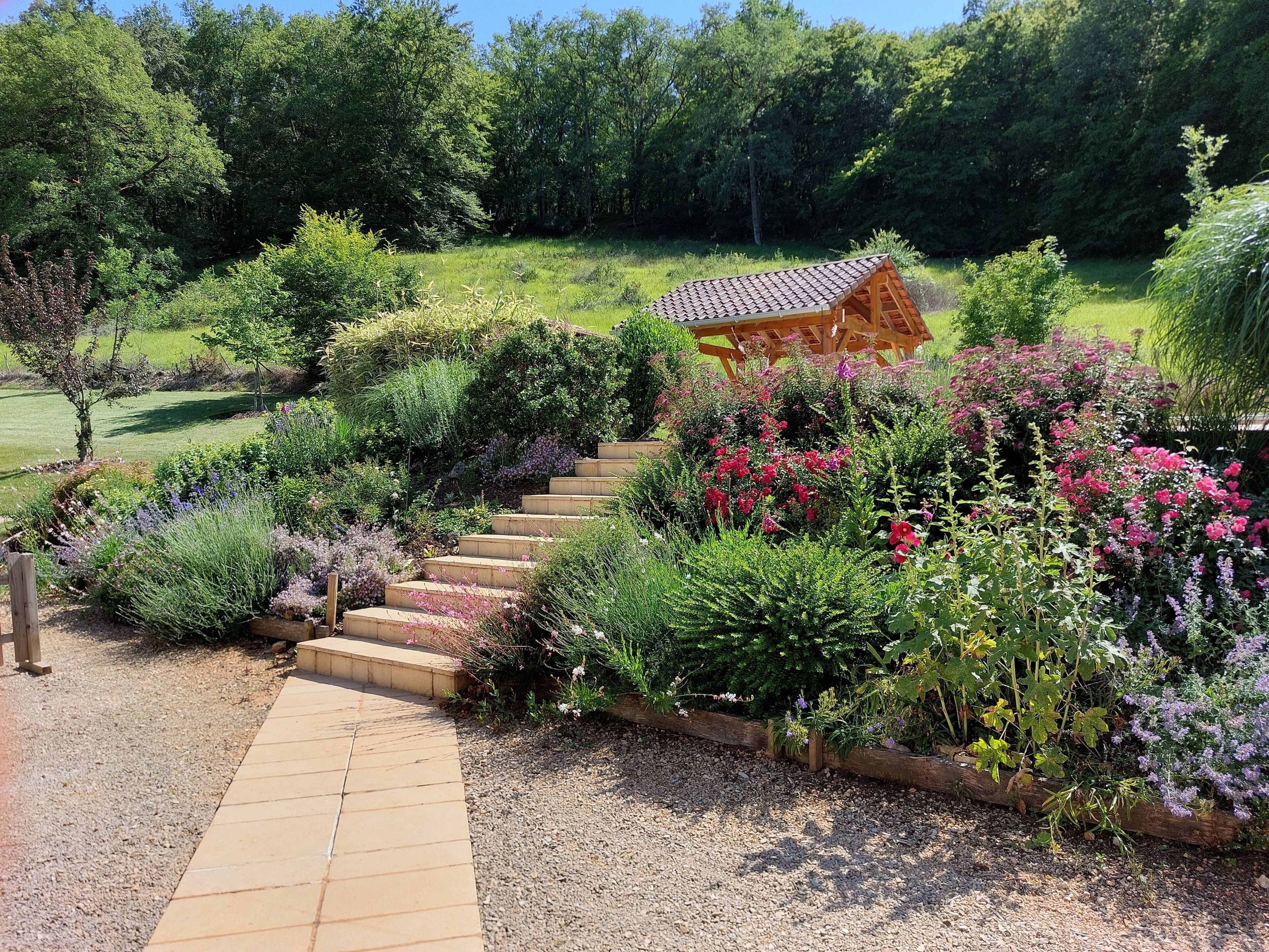 Ein Garten mit einer Holzpergola, einem Kiesweg und verschiedenen Pflanzen.