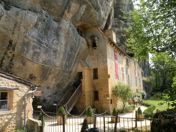 A stone building with a staircase leading up to a window, surrounded by greenery and a fenced area.