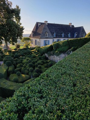 A large, traditional house with a steeply sloped roof, surrounded by meticulously maintained hedges and trees.