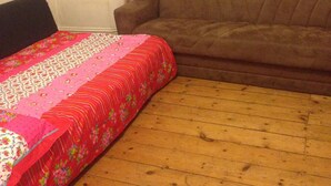 Une pièce avec un plancher en bois, un canapé marron, un lit recouvert d’une courtepointe à motifs floraux et un rideau.