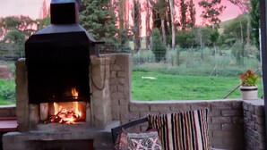An outdoor stone fireplace with a fire, surrounded by a stone wall and a grassy area with trees in the background.