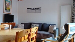 Ett vardagsrum med ett matbord i trä, två trästolar, en grå soffa och en tv-apparat placerad på ett träställ.