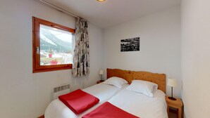 A bedroom with a wooden headboard, two beds, a window with a view of mountains, a heater, and a small bedside table with a lamp. - Balcons G 025 - PARC NAT. VANOISE appart. 8 pers. (Val Cenis)
