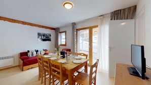 A dining area with a wooden table set for four, a sofa with red cushions, a television on a stand, and a window with curtains. - Balcons G 025 - PARC NAT. VANOISE appart. 8 pers. (Val Cenis)