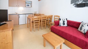 Ein gemütlicher Wohnraum mit Küchenzeile, einer Essecke mit Holzstühlen, einem roten Sofa mit gemusterten Kissen und einem Fernseher auf einem Holzboden.