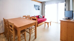 Ein Zimmer mit Esstisch und Stühlen aus Holz, einem roten Sofa mit Kissen, einem kleinen Holztisch mit Computer und einem Fenster mit Vorhängen.
