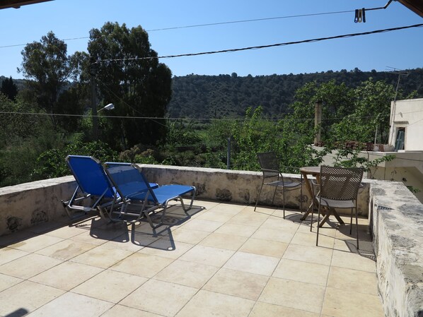 A terrace with blue lounge chairs and a table, overlooking a lush landscape with trees and hills.
